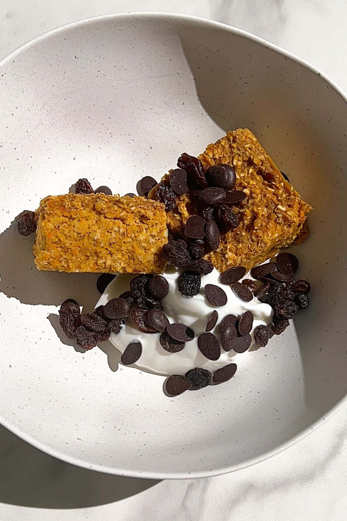 Carrot Cake Baked Oatmeal Bars in a bowl with yogurt, raisins and chocolate chips.