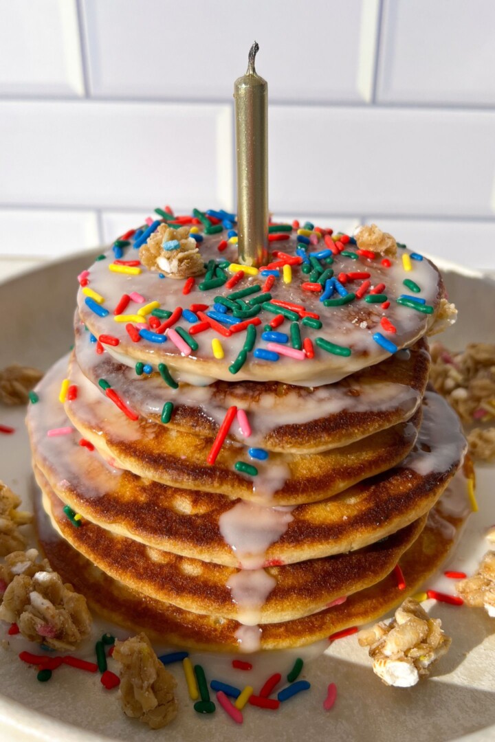 Stack of birthday cake pancakes with toppings and a candle in the middle.