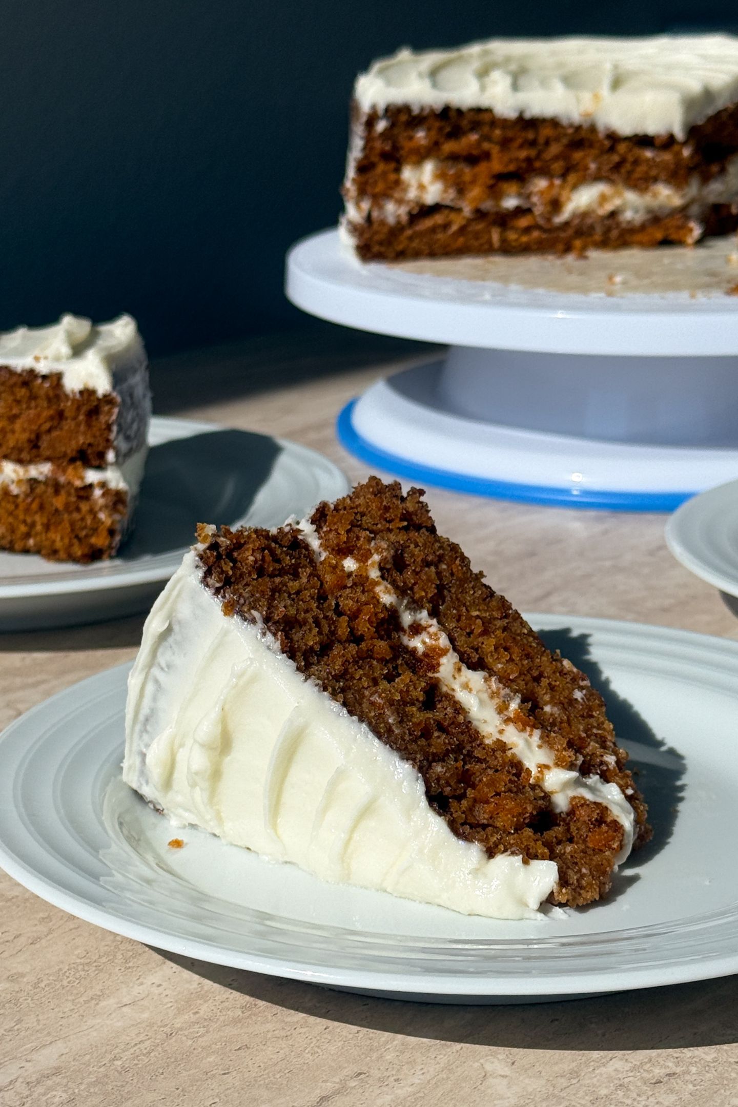 Slice of carrot cake on a plate with the rest of the cake in the background.
