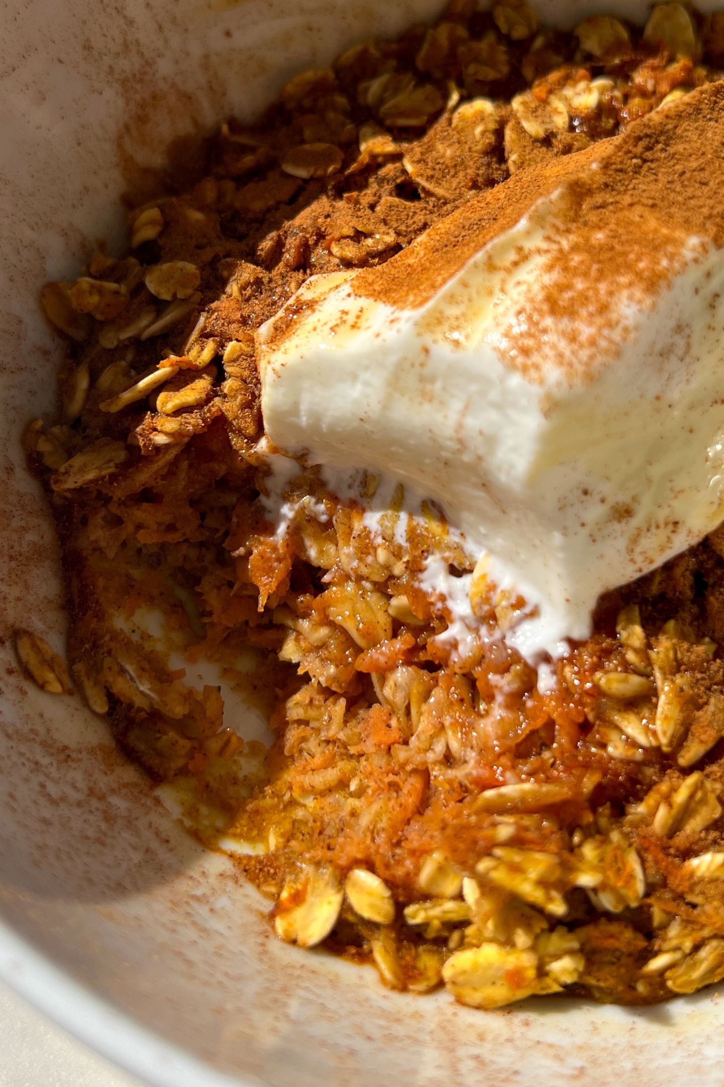 Carrot cake oatmeal in a bowl topped with yogurt, cinnamon and maple syrup with a scoop taken out.