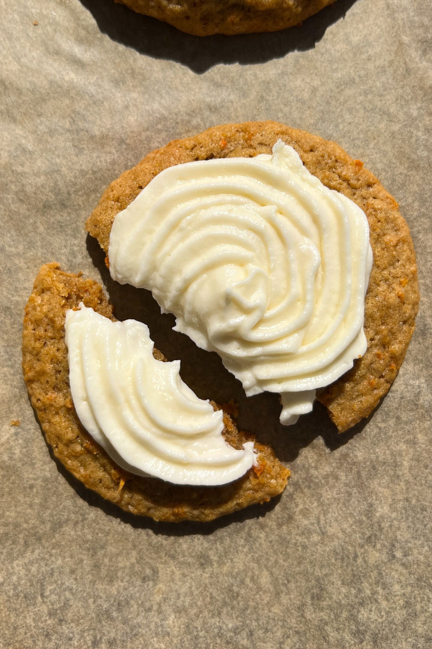 A Frosted Carrot Cookie broken in half on a baking sheet that is lined with parchment paper.