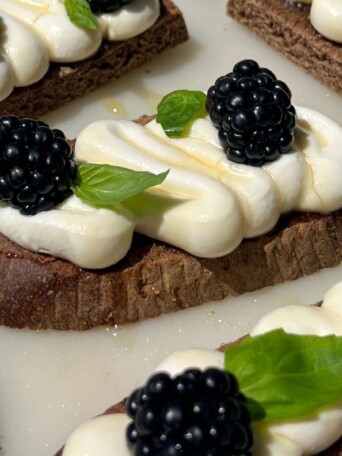 A piece of pumpernickel crostini topped with goat cheese, honey, fresh basil and blackberries.