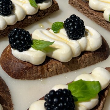 A piece of pumpernickel crostini topped with goat cheese, honey, fresh basil and blackberries.