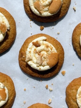 Banana Pudding Cookies topped with frosting and crushed wafer cookies on parchment paper.
