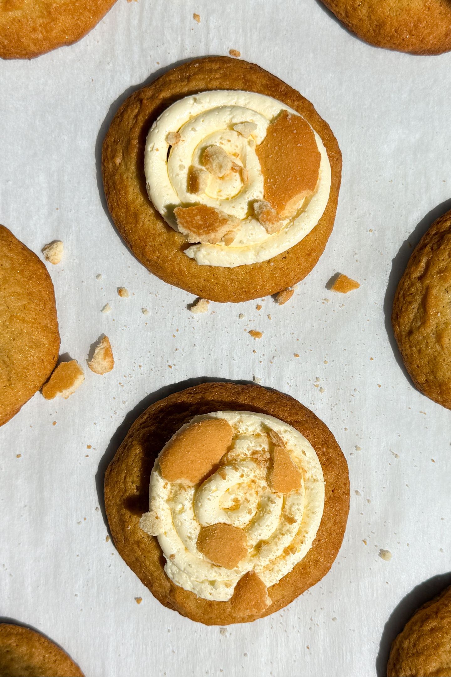 Banana pudding cookies on parchment paper topped with a banana buttercream frosting and crushed vanilla wafer cookies.